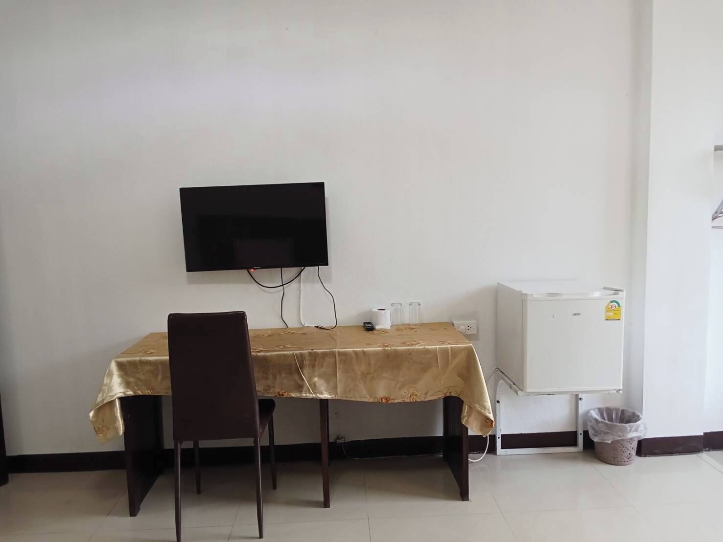 A small room with a table and chair, a television mounted on the wall, a small refrigerator, and a trash bin.