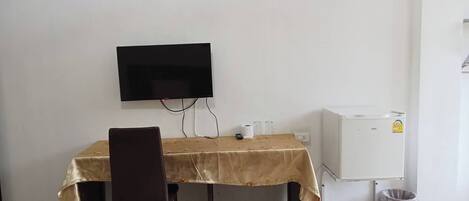 A small room with a table and chair, a television mounted on the wall, a small refrigerator, and a trash bin.