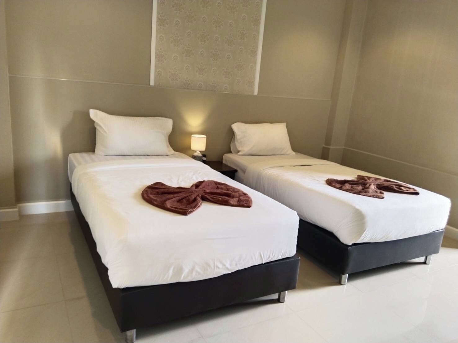 Dos camas individuales con sábanas blancas y toallas marrones dobladas encima, una mesita de noche con lámpara, y una pared lisa con un panel decorativo.