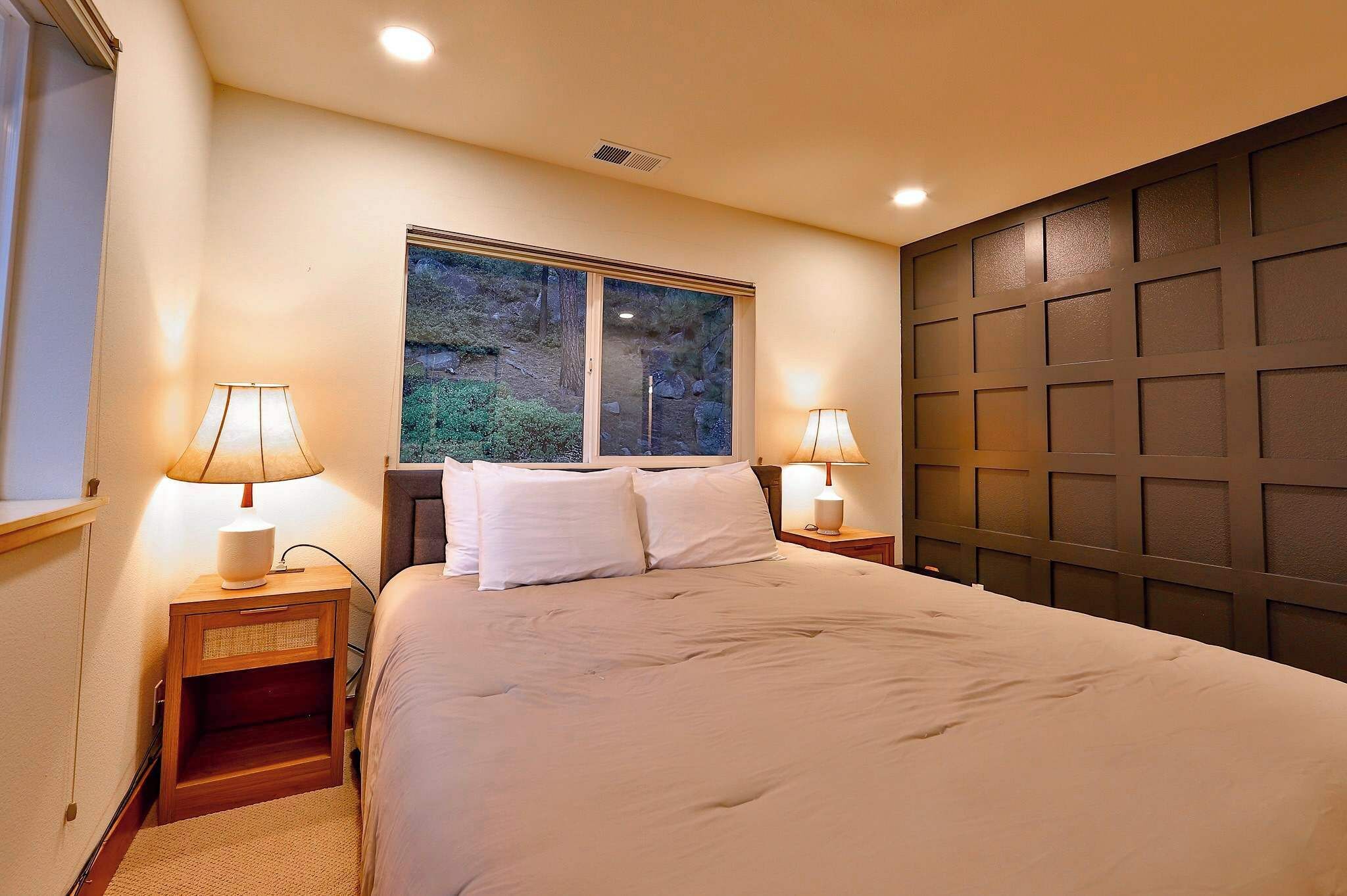 A bedroom with a large bed, two bedside lamps, a window with a view of greenery, and a dark headboard.