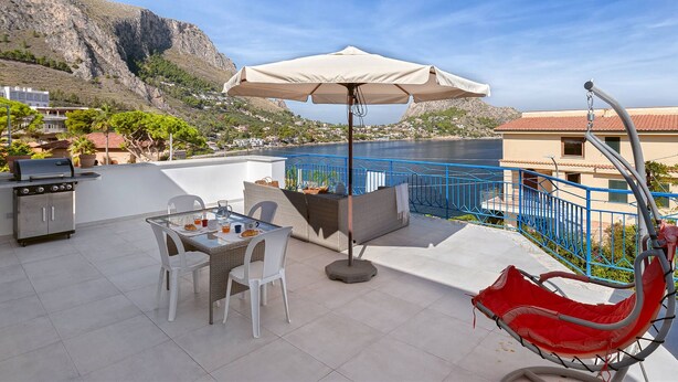 A terrace with a table, chairs, a grill, and a view of the sea and mountains.