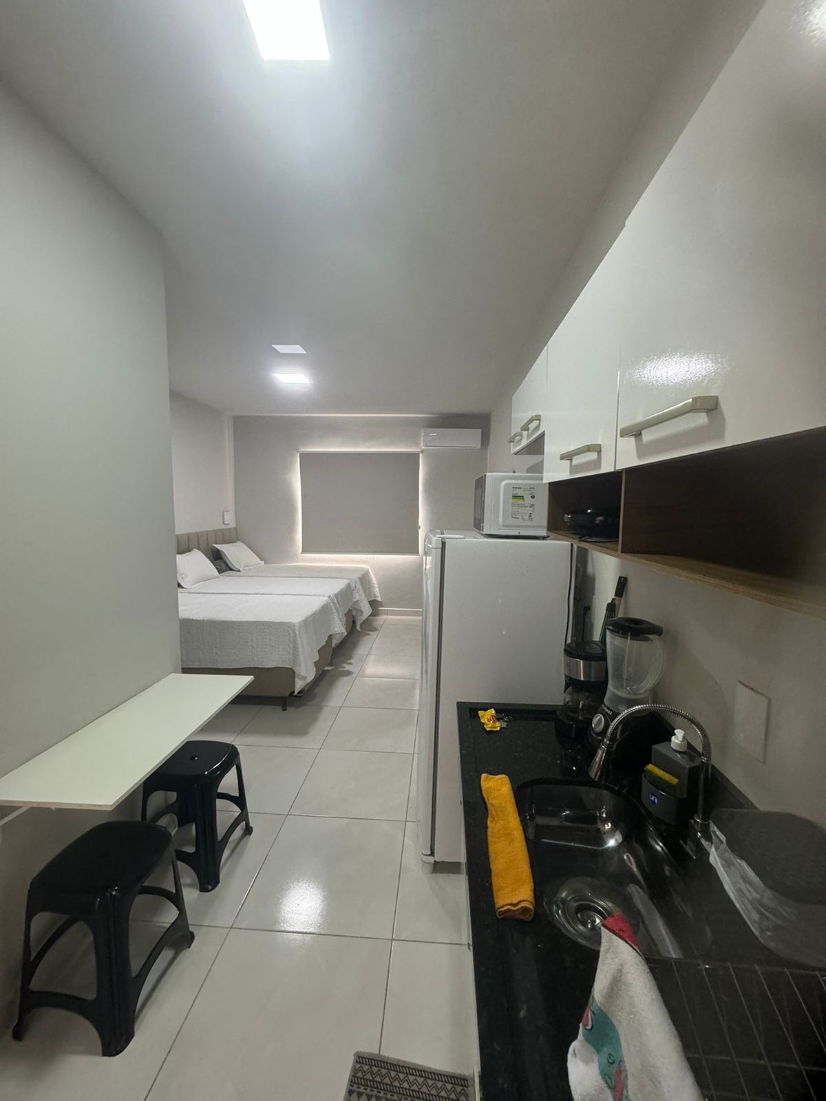 Habitación de hotel compacta con kitchenette, una cama con cubrecama blanco, una mesita con dos taburetes negros y un microondas en la encimera.