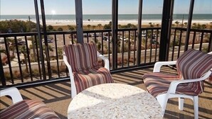Un balcon avec des chaises à rayures et une table ronde, donnant sur une plage et un parking.
