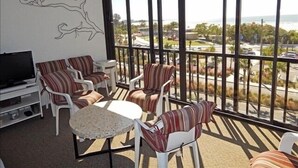 Un balcon avec des chaises blanches et une petite table, donnant sur un quartier résidentiel bordé de palmiers et traversé par une route.