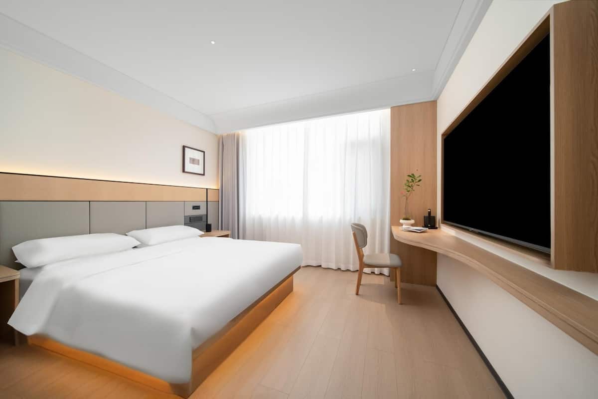A modern hotel room with a large bed, a desk with a flat-screen TV, a chair, and a window with sheer curtains.