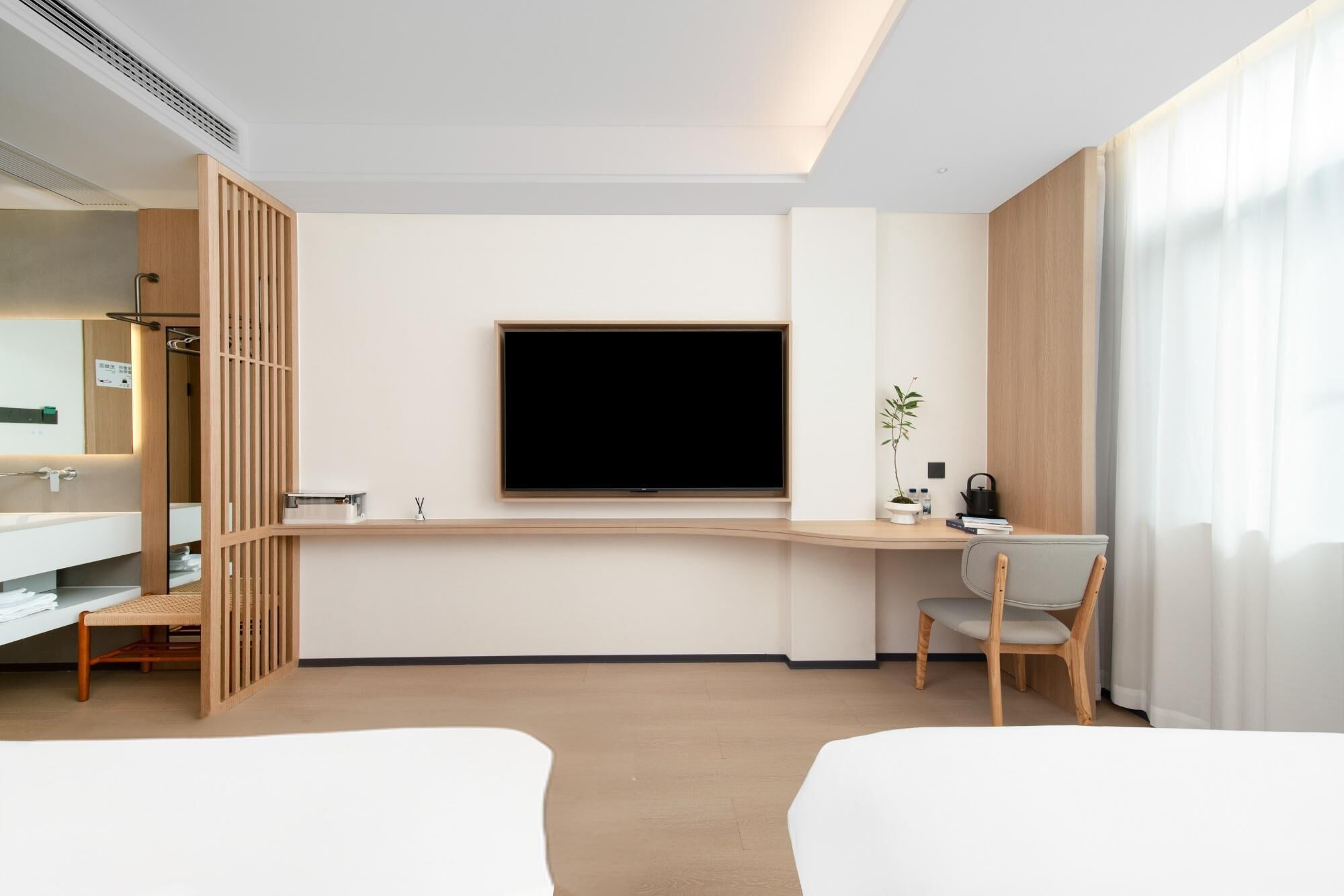 A modern hotel room with a large flat-screen TV, a wooden desk, a chair, a bed, and a bathroom with a mirror and sink.
