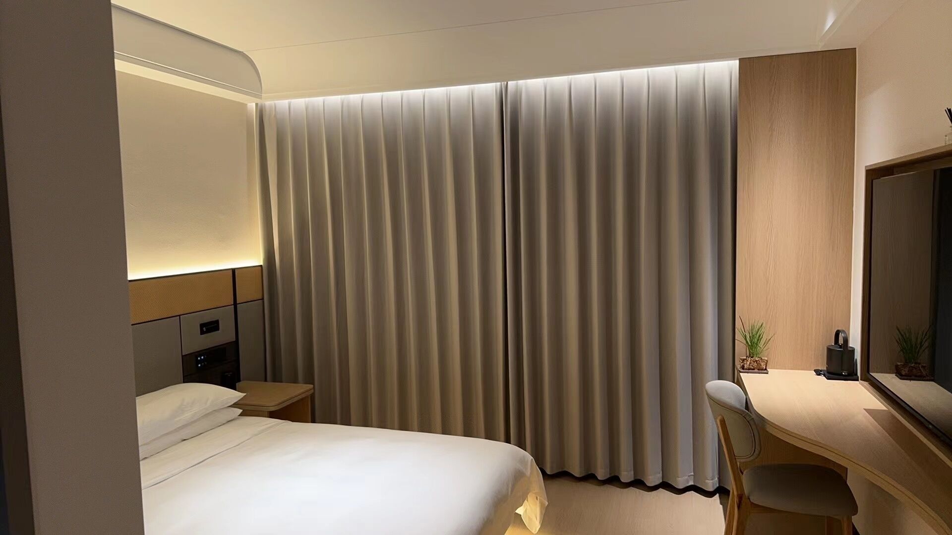 A hotel room with a bed, a desk, a chair, a nightstand, a lamp, and a large window with curtains.