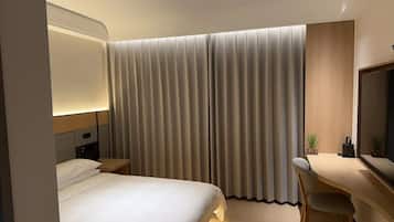 A hotel room with a bed, a desk, a chair, a nightstand, a lamp, and a large window with curtains.