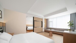 Ein modernes Hotelzimmer mit einem großen Bett, einer kleinen Sitzecke mit Tisch, einem Schreibtisch mit Stuhl und einem großen Fenster mit Vorhängen.