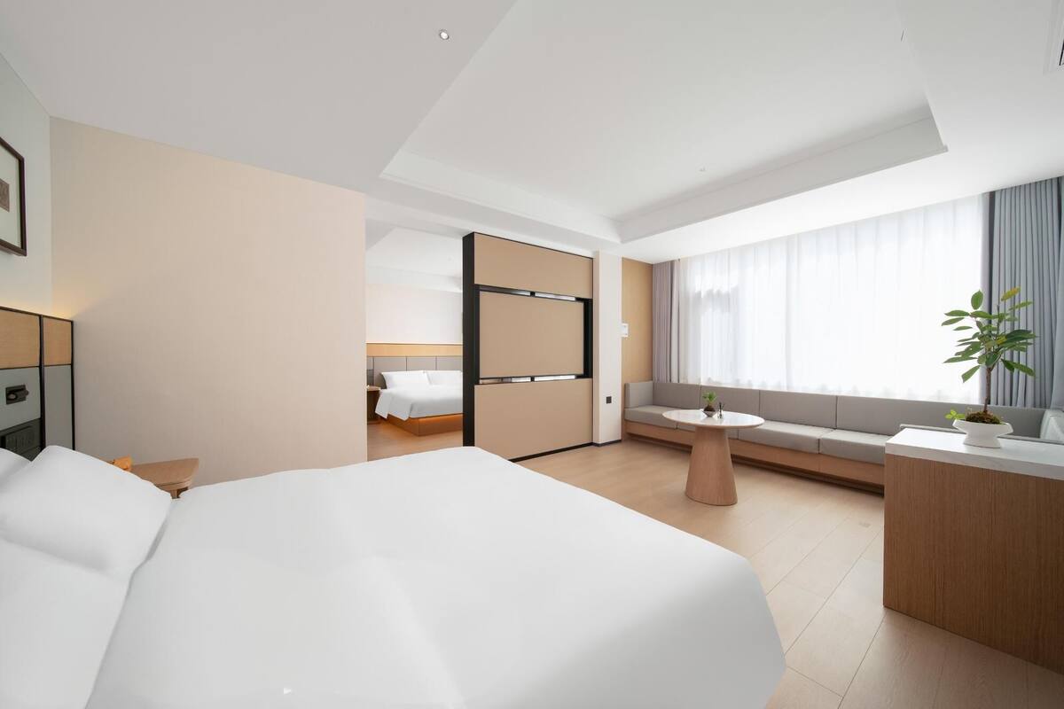A modern hotel room with a large bed, a small seating area with a table, a desk with a chair, and a large window with curtains.