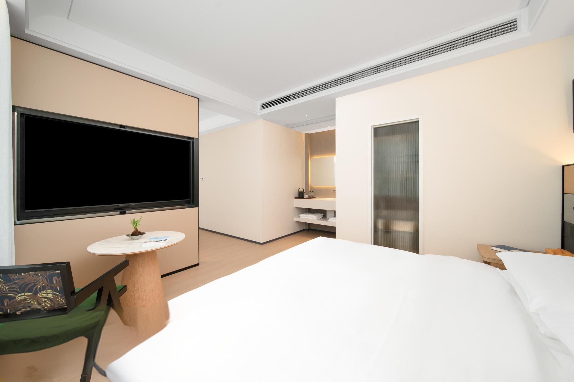 A modern hotel room with a large bed, a flat-screen TV, a small round table with a plant, a green chair, and a walk-in closet with a mirror and shelves.