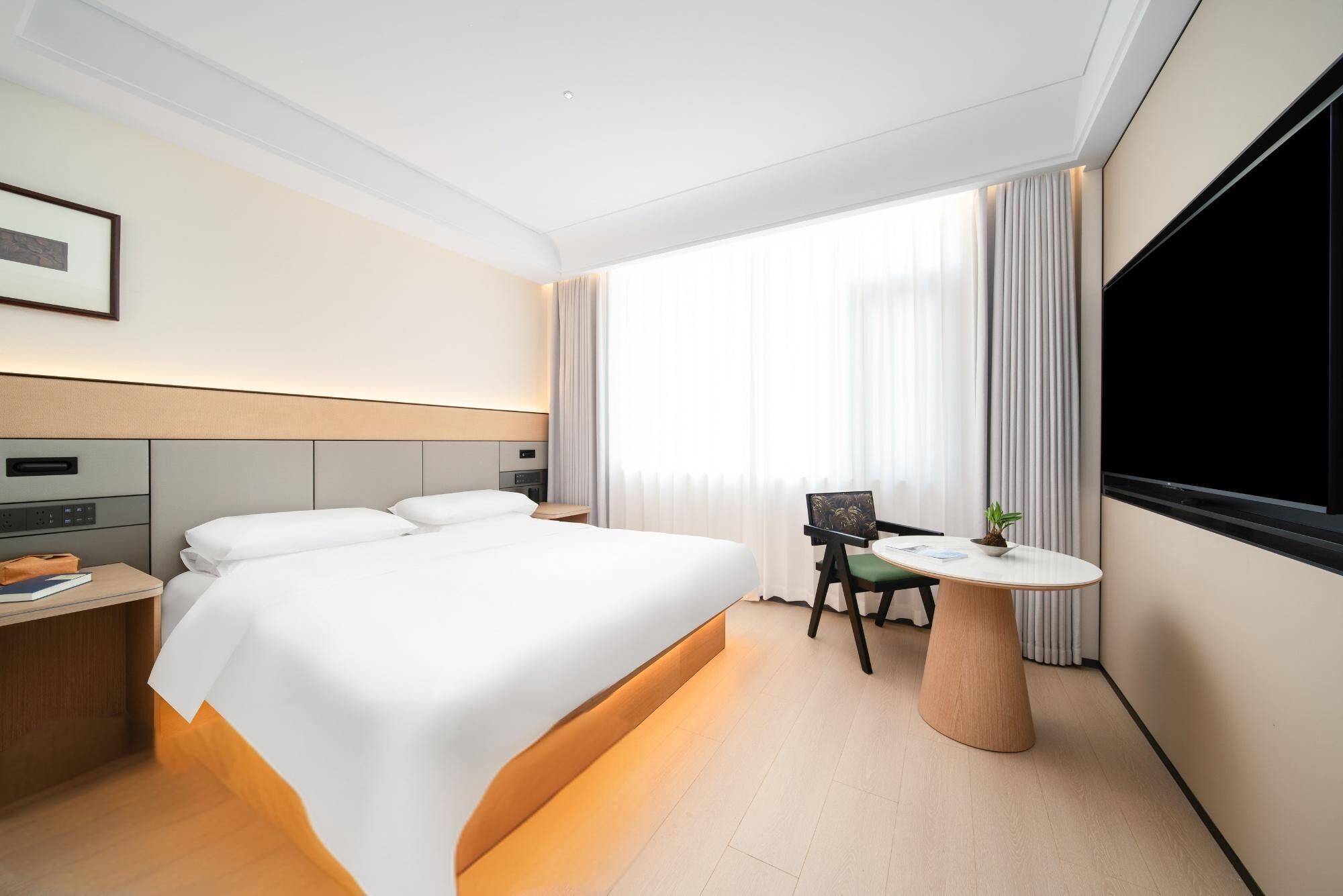 A modern hotel room with a large bed, a desk with a chair, a flat-screen TV, and a large window with curtains.