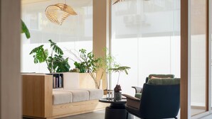 Ein modernes Wohnzimmer mit einer Couch, einem Sessel, einem Couchtisch und einem Bücherregal. Große Fenster und hängende Korbtabletten sind vorhanden.
