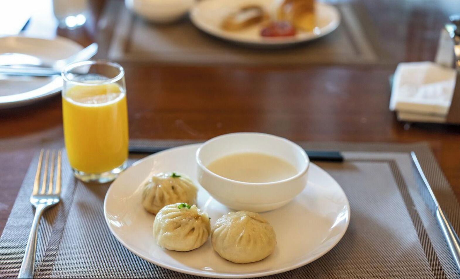 Un plato con dumplings y un tazón de sopa, un vaso de jugo de naranja y un tenedor sobre una mesa.