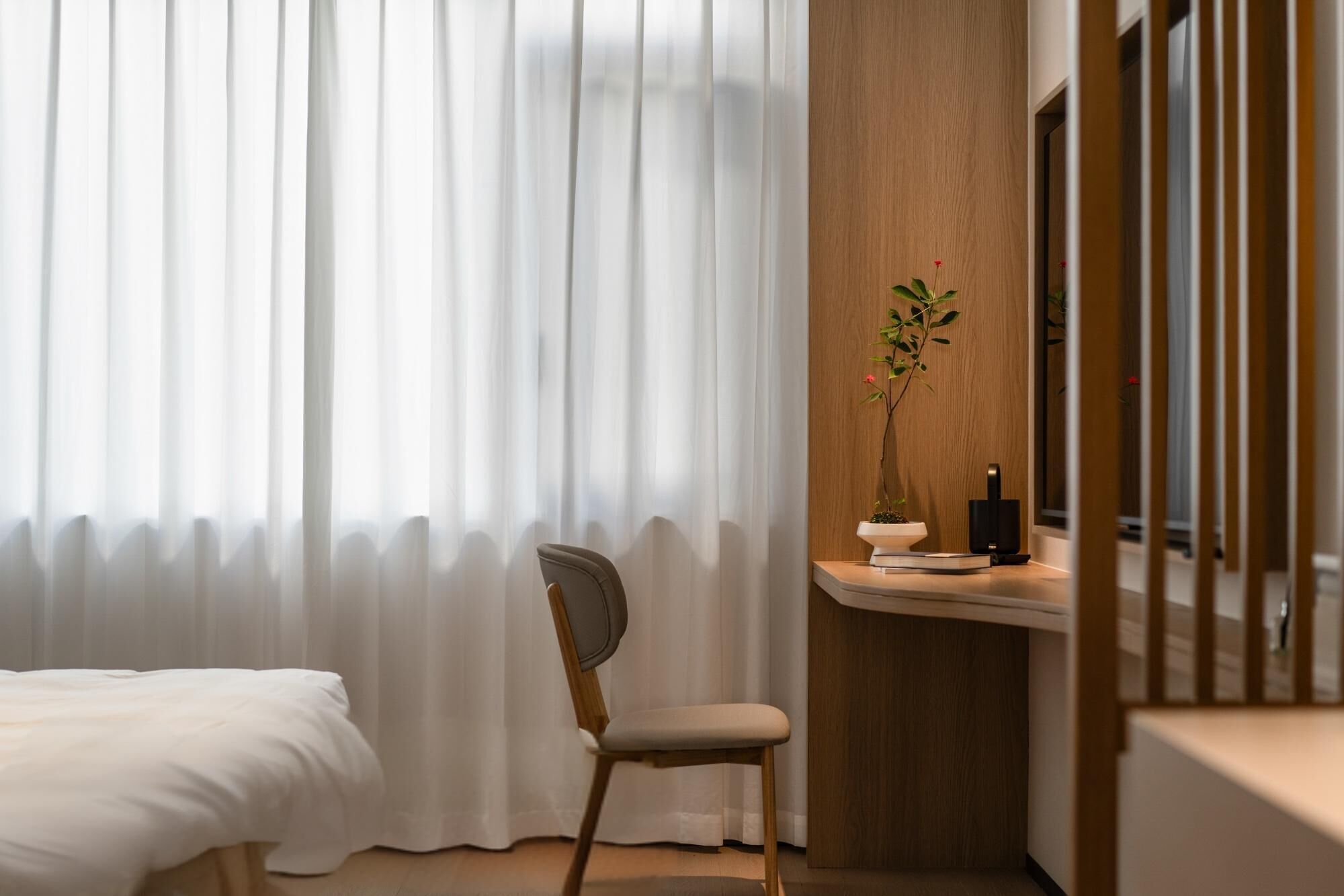A hotel room with a bed, a chair, a wooden desk with a plant, and a window with sheer curtains.