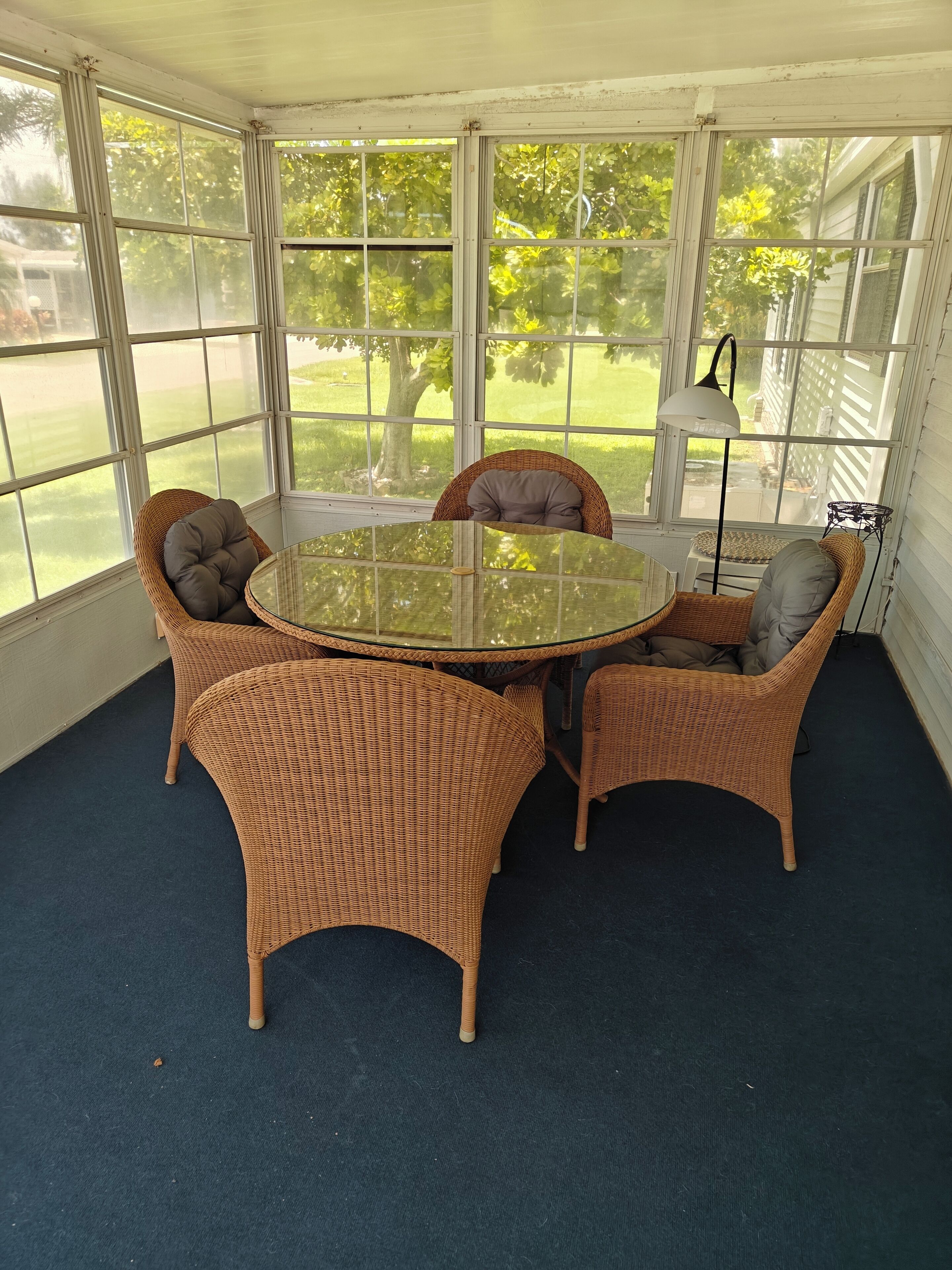 Un porche cubierto con una mesa redonda de vidrio rodeada de sillas de mimbre, ventanas amplias y una lámpara.