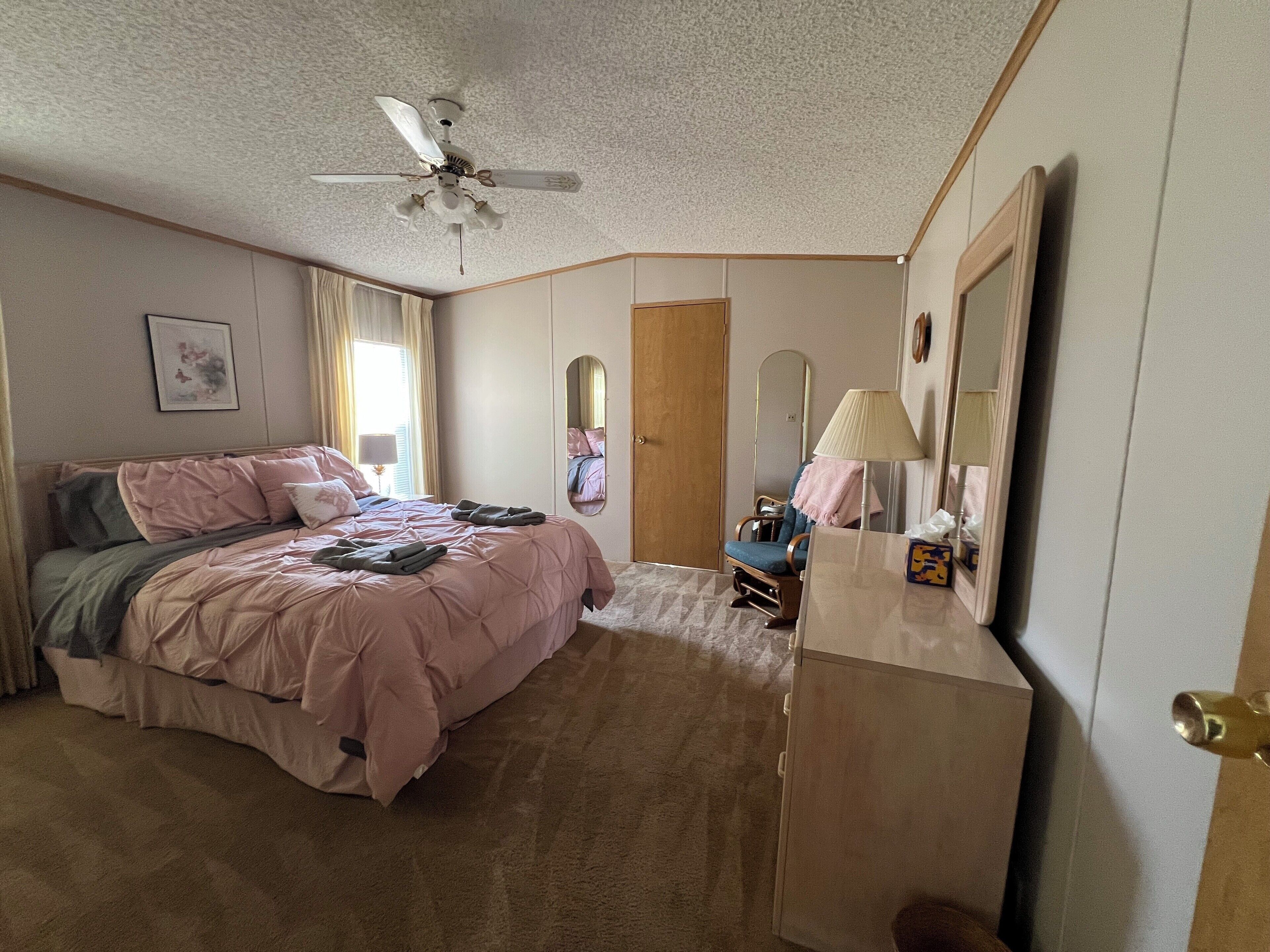 Un dormitorio con una cama, un ventilador de techo, un aparador, un espejo y una puerta.