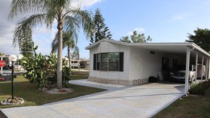 A single-story mobile home with a covered porch, a driveway, and a palm tree in the foreground. - Golfing, swimming and a friendly secure community (55+) - SEASONAL (30days+) (North Fort Myers)