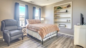 Une chambre à coucher comprenant un lit, une chaise, un ventilateur de plafond, une fenêtre avec des rideaux, une étagère avec des objets décoratifs et un téléviseur.