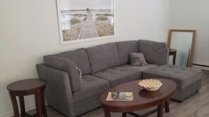 A modern living room with a grey sectional sofa, a round coffee table, and a small wooden side table. A framed picture of a pier is on the wall.