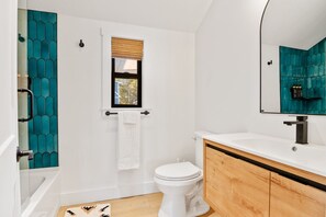 A modern bathroom with a white toilet, a wooden vanity, a large mirror, and teal-patterned tiles on the walls.