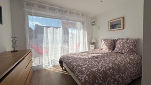 Un dormitorio con cama, cómoda de madera, mesita de noche, ventana con cortinas traslúcidas y vistas al exterior.