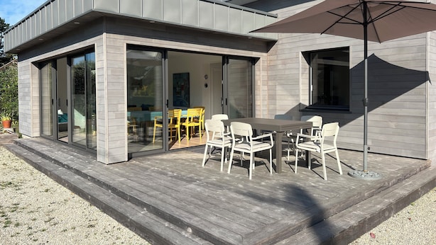 Una casa moderna con un patio amoblado, que incluye una mesa, sillas, un gran paraguas y un ventanal corredero que da a un espacio interior.