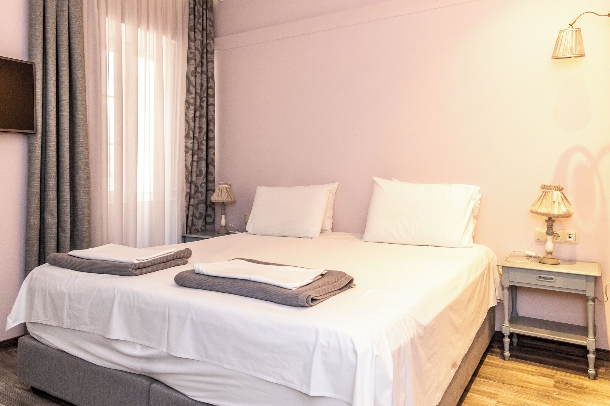 A neatly made bed with white and grey linens, a bedside table with a lamp, and a wall-mounted TV.