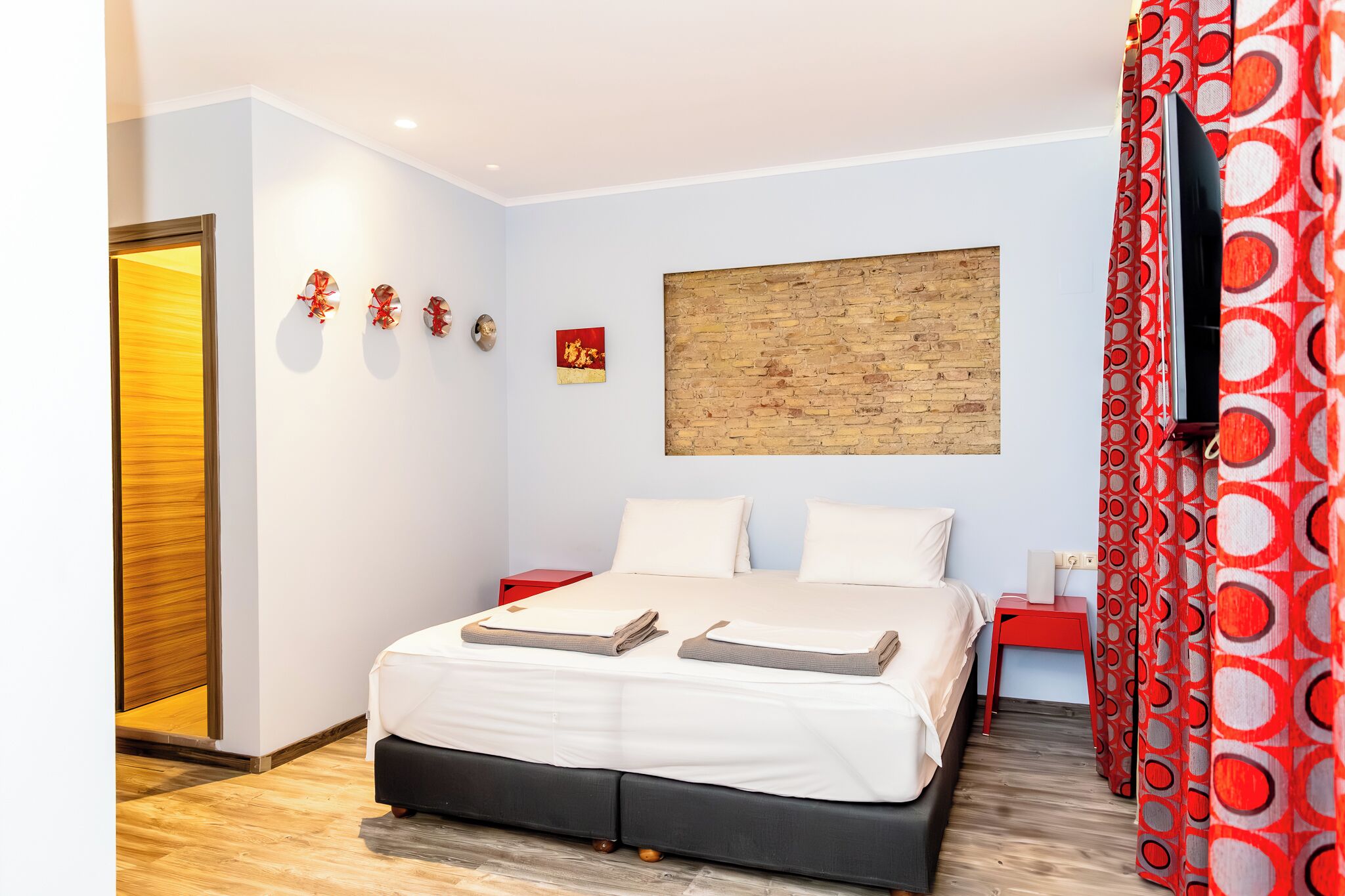 A modern hotel room with a bed, a wooden headboard, a red side table, and a large window.