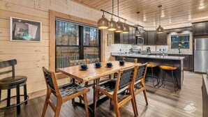 Een houten interieur met een eettafel, stoelen, een keukenhoek en hangende hanglampen.