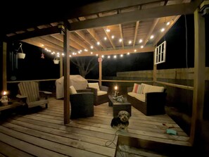 An outdoor patio with wooden flooring, covered seating, string lights, and a small dog.
