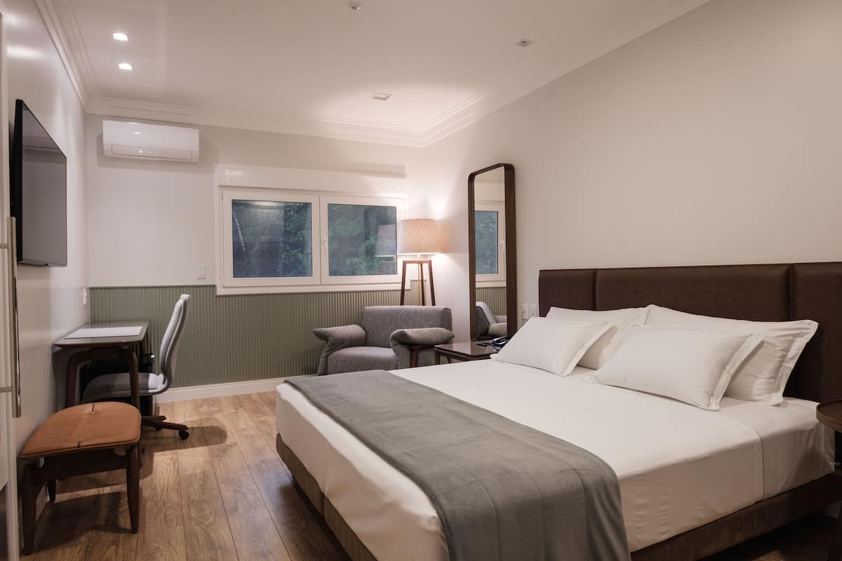 A hotel room with a large bed, a desk, a chair, a mirror, and a window with a view of trees.
