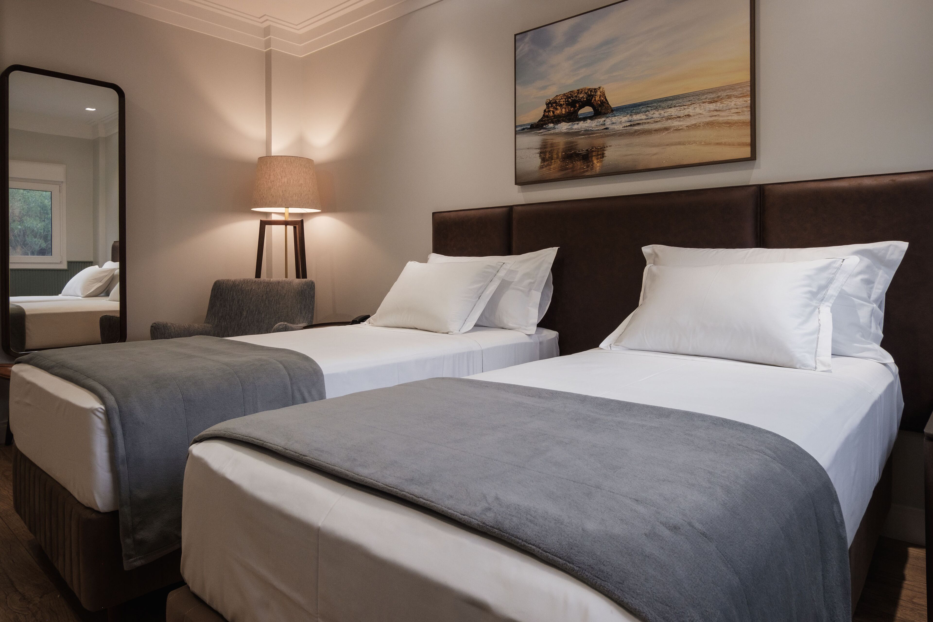 A hotel room with two beds, a large mirror, a lamp, and a painting of a bridge over water.