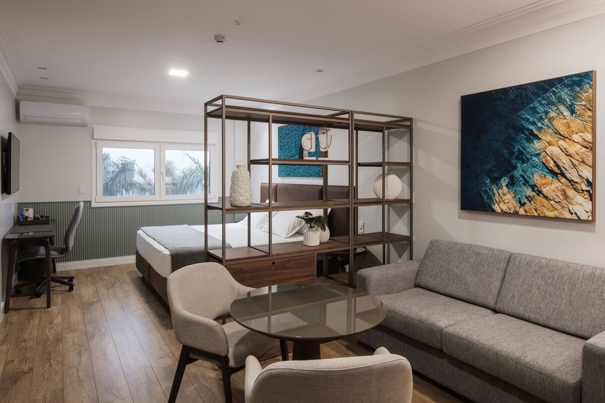A modern hotel room with a bed, a sofa, a glass-top table, a desk, and a large abstract painting on the wall.