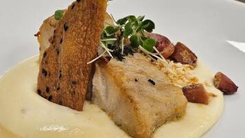 A dish with a piece of fish, bread, and greens on a white plate.