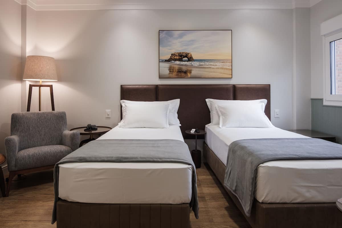 A hotel room with two beds, a chair, a lamp, and a painting of a coastal scene.