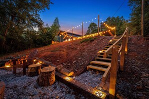 Una escalera de madera lleva a una cabaña con luces de cuerda, rodeada de árboles y grava.