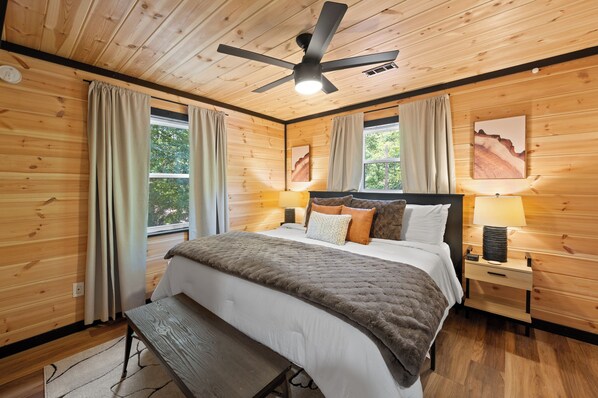 Un dormitorio con paneles de madera, una cama, dos mesitas de noche con lámparas, un ventilador de techo y una ventana con cortinas.