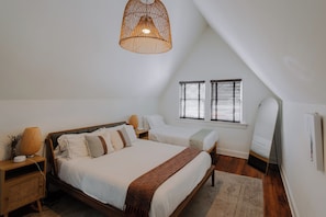 A bedroom with a large bed, bedside tables, a hanging light fixture, a window with blinds, and a mirror.