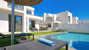 Un espace piscine extérieur moderne, doté de chaises longues, d’une table et d’un parasol en chaume.