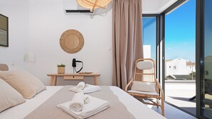 Une chambre d’hôtel moderne avec un grand lit, une table de chevet en bois, un fauteuil en osier et une vue sur la ville grâce à de grandes fenêtres.
