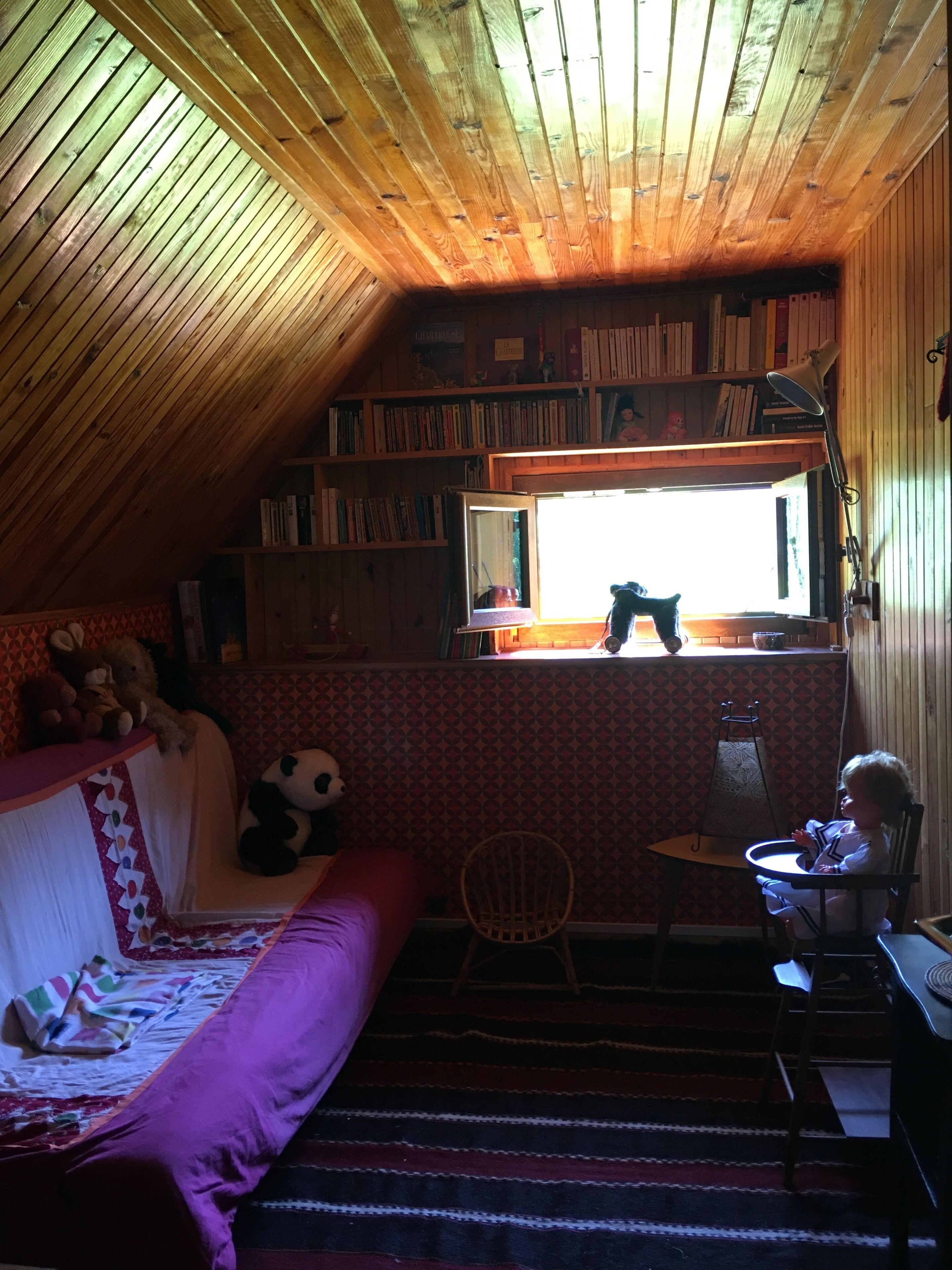 A cozy attic bedroom with a sloped ceiling, a large window, a bookshelf, a bed with a colorful quilt, a chair, and a small table.