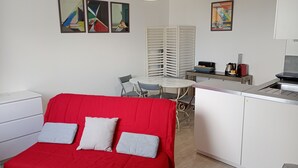 A compact room with a red sofa, a small dining table with chairs, a kitchenette, and artwork on the walls.