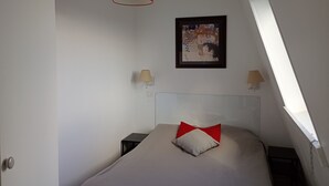 A bedroom with a bed, a red pendant light, two bedside lamps, a framed artwork, and a skylight.
