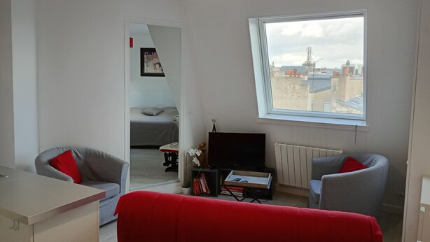 A modern living room with a sofa, a TV, a window with a city view, and a door leading to a bedroom.