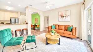 A modern living room with a sofa, green chairs, a coffee table, and a kitchen in the background. - Upstay - Condo w Hot Tub and Pool - Near Disney (Winter Garden)