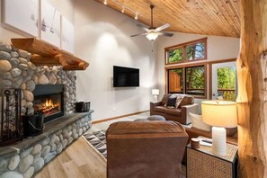 A cozy living room with a stone fireplace, a ceiling fan, and a large window with a view of trees.
