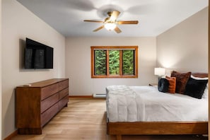 A bedroom with a wooden bed, a ceiling fan, a TV mounted on the wall, and a window with a view of trees.