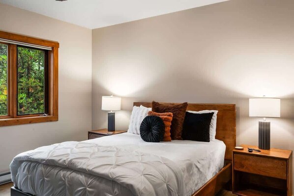 A bedroom with a large bed, two bedside tables, and a window with a view of trees.