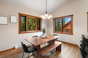 Un comedor con mesa de madera, sillas negras y un banco. Se observan ventanas grandes con marcos de madera y una lámpara colgante.